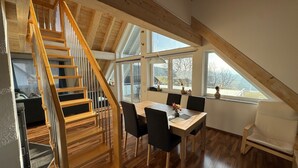 A spacious room with a wooden staircase, a dining area with a table and chairs, and large windows offering a view of the outdoors. - 5-star maisonette vacation apartment Maria with fantastic Black Forest views (Schramberg)