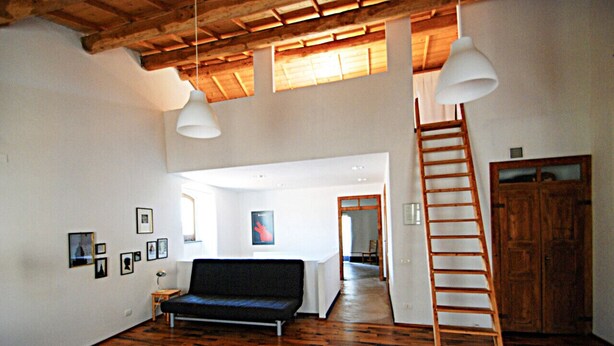 Um quarto amplo com piso de madeira, um sofá preto, uma escada e uma porta branca.