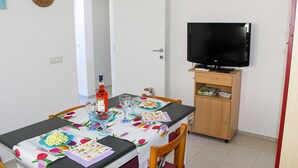 Uma área de refeições com uma mesa posta para dois, uma televisão sobre um suporte de madeira e uma porta que dá para outro quarto.