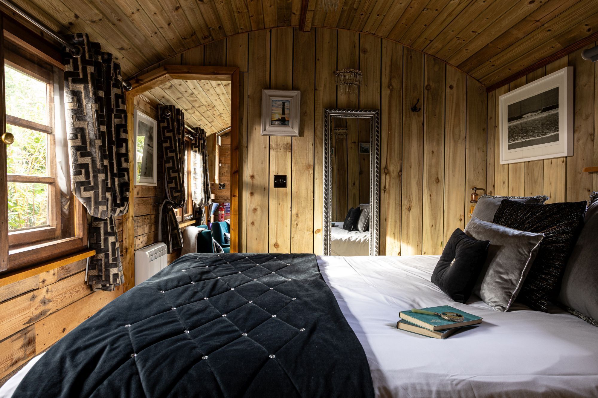A cozy cabin-style bedroom with a large bed, a window with curtains, a door leading to another room, and a framed picture on the wall.