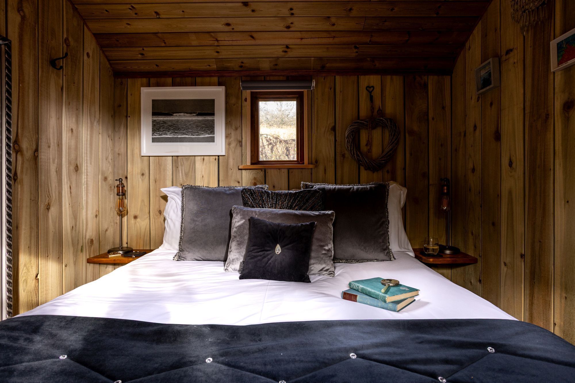 A wooden cabin bedroom with a bed, pillows, a small table with books, a window, and a framed picture on the wall.