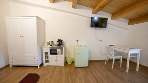 Een ruime, goed verlichte kamer met een houten plafond, een witte kledingkast, een kitchenette met een magnetron en koelkast, een tv aan de muur en een wit bureau met een stoel.
