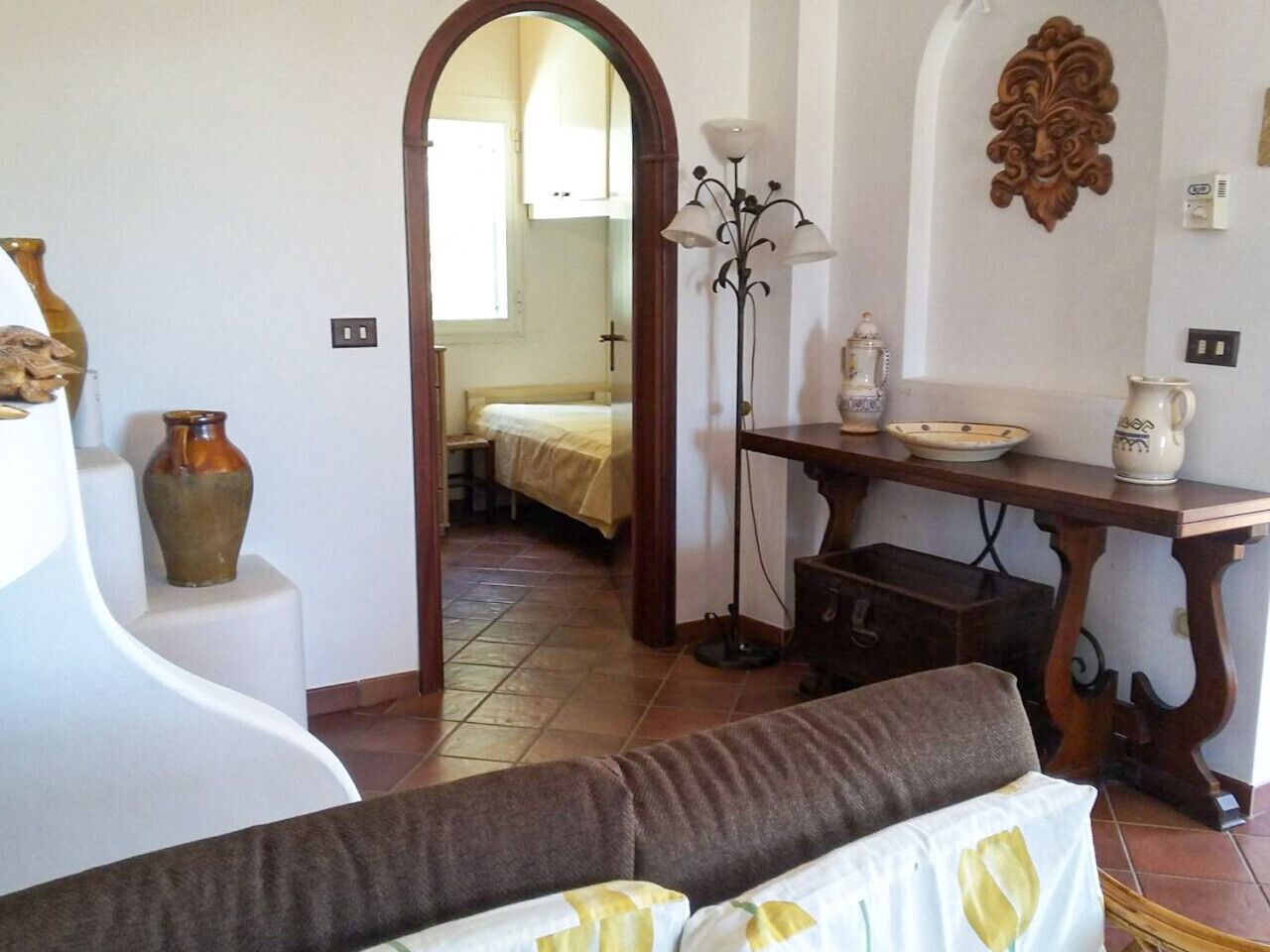 A room with a sofa, a table with decorative items, a floor lamp, and a doorway leading to another room with a bed.
