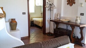 Een kamer met een bank, een tafel met decoratieve objecten, een vloerlamp en een deuropening naar een andere kamer met een bed.