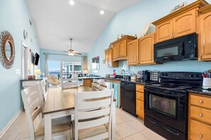 A modern kitchen with wooden cabinets, a black stove, and a dining area with a table and chairs. - 🌞🌴Baywatch G9 | Water Views + Walk to Beach | Handicap Accessible (Pensacola Beach)