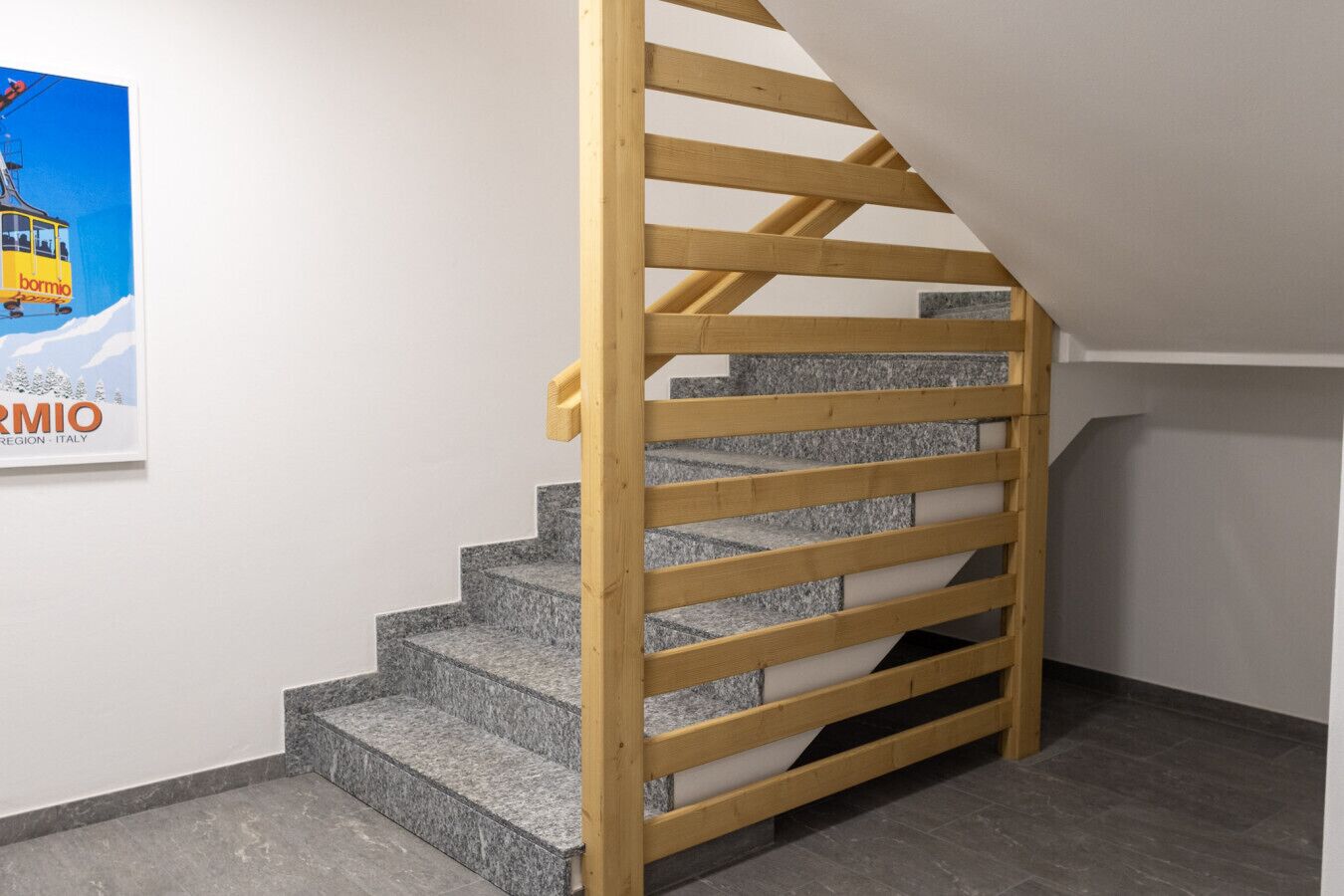 Een trap met houten leuningen en granieten treden, die leidt naar een plateau met een witte muur en een poster.