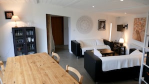 Un salon moderne avec une table à manger en bois, des canapés noirs et un divan blanc. On y trouve un meuble contenant de la verrerie et un téléviseur fixé au mur.
