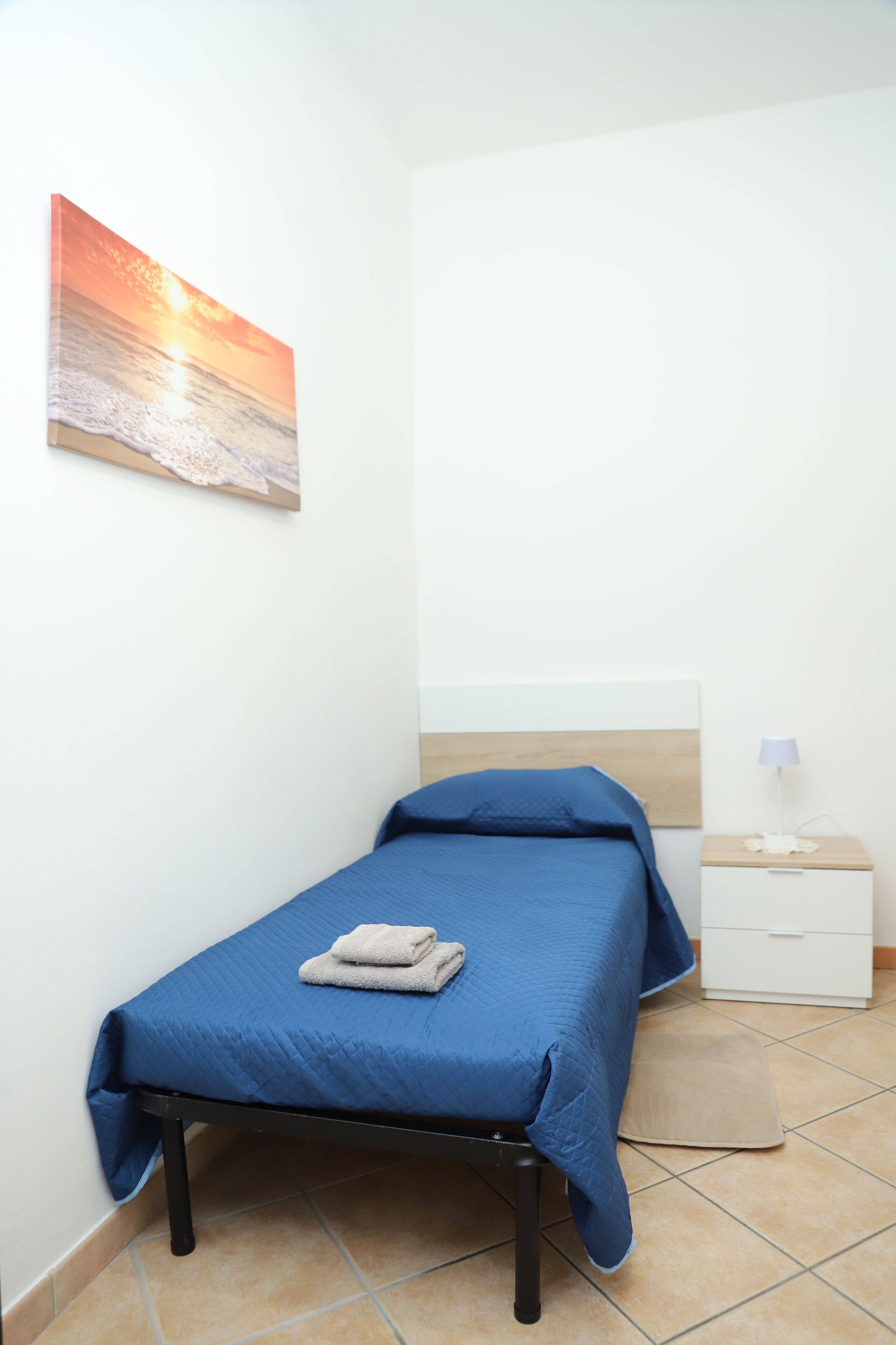 A small, neatly arranged bedroom with a single bed, a bedside table with a lamp, and a wall-mounted painting of a sunset over the ocean.