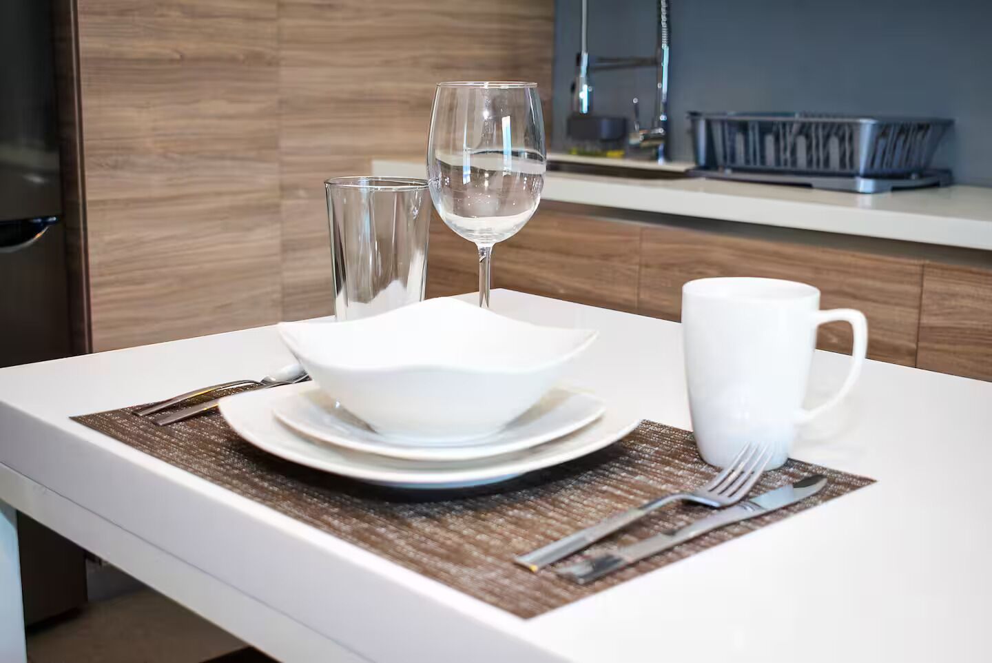 Een moderne keuken met een wit aanrecht, een witte kom, een glas water, een witte mok en een vork op een onderzetter.