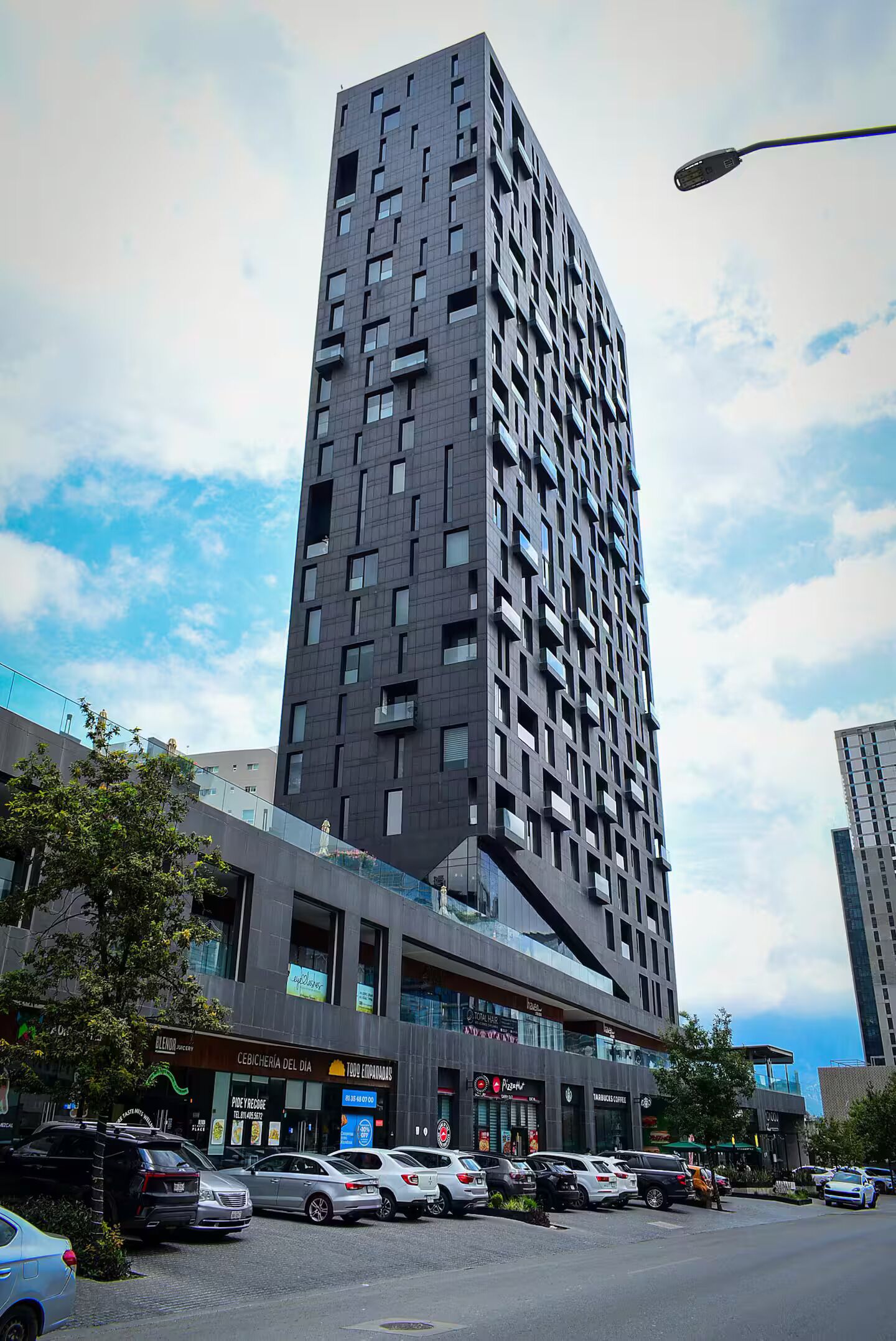 A tall, modern building with a grid-like facade, multiple balconies, and a ground-level commercial area with shops and parked cars.