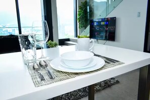 Mesa de comedor moderna con platos blancos, una copa y cubiertos, con vista a la ciudad a través de amplias ventanas.