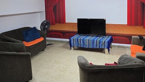 Una sala de estar con un sofá, sillones, una mesa de centro, un televisor y un área de escenario con cortinas rojas.