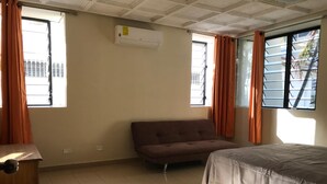 Un dormitorio con cama, sofá y escritorio. Hay dos ventanas con cortinas y un equipo de aire acondicionado en la pared.