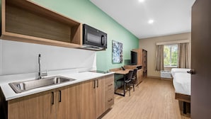 A kitchenette with a microwave, sink, and wooden cabinets. There is a desk with a chair and a bed with curtains. - Echo Suites Extended Stay By Wyndham Bozeman (Bozeman)