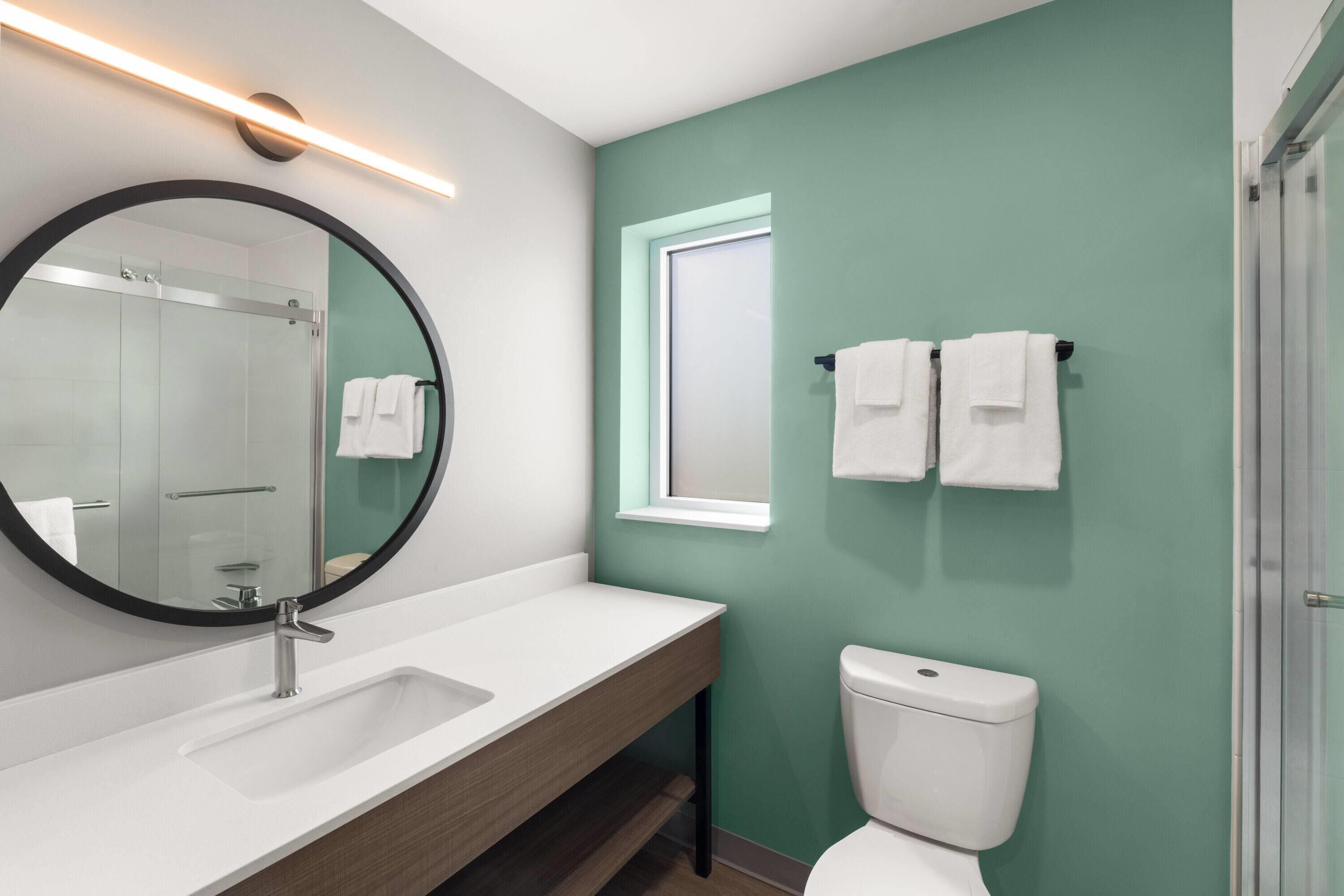 Une salle de bain avec un lavabo blanc, un miroir rond, des toilettes et un espace douche.