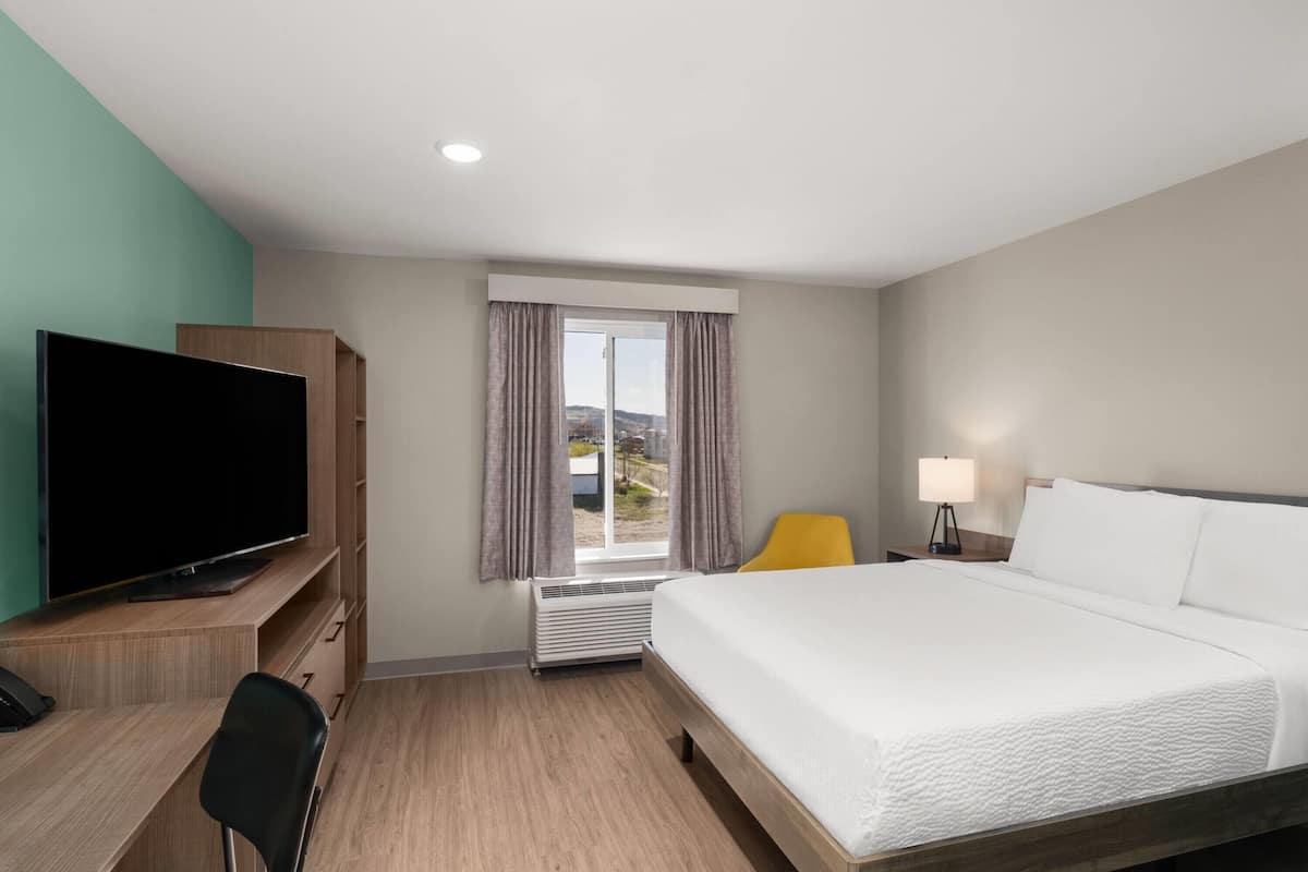 Una habitación de hotel con cama, escritorio con televisor, silla, lámpara y ventana con cortinas.