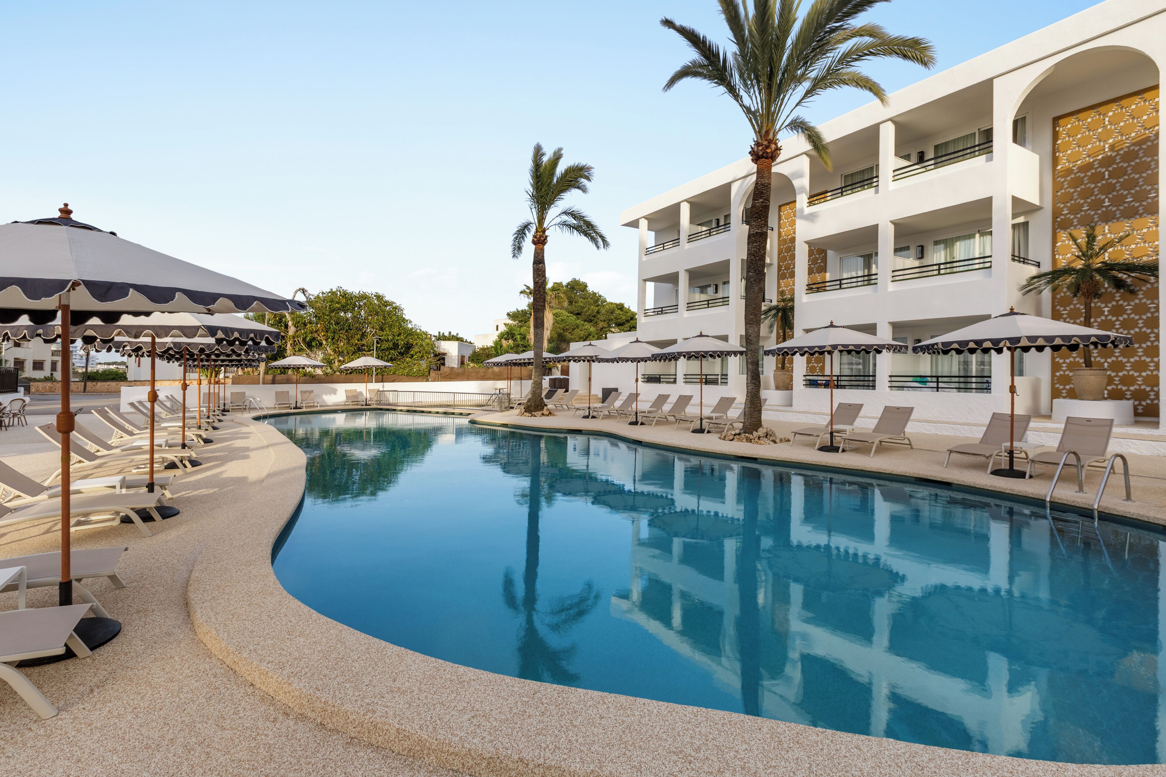 A pool area with lounge chairs and umbrellas, a multi-story building with balconies, and palm trees.