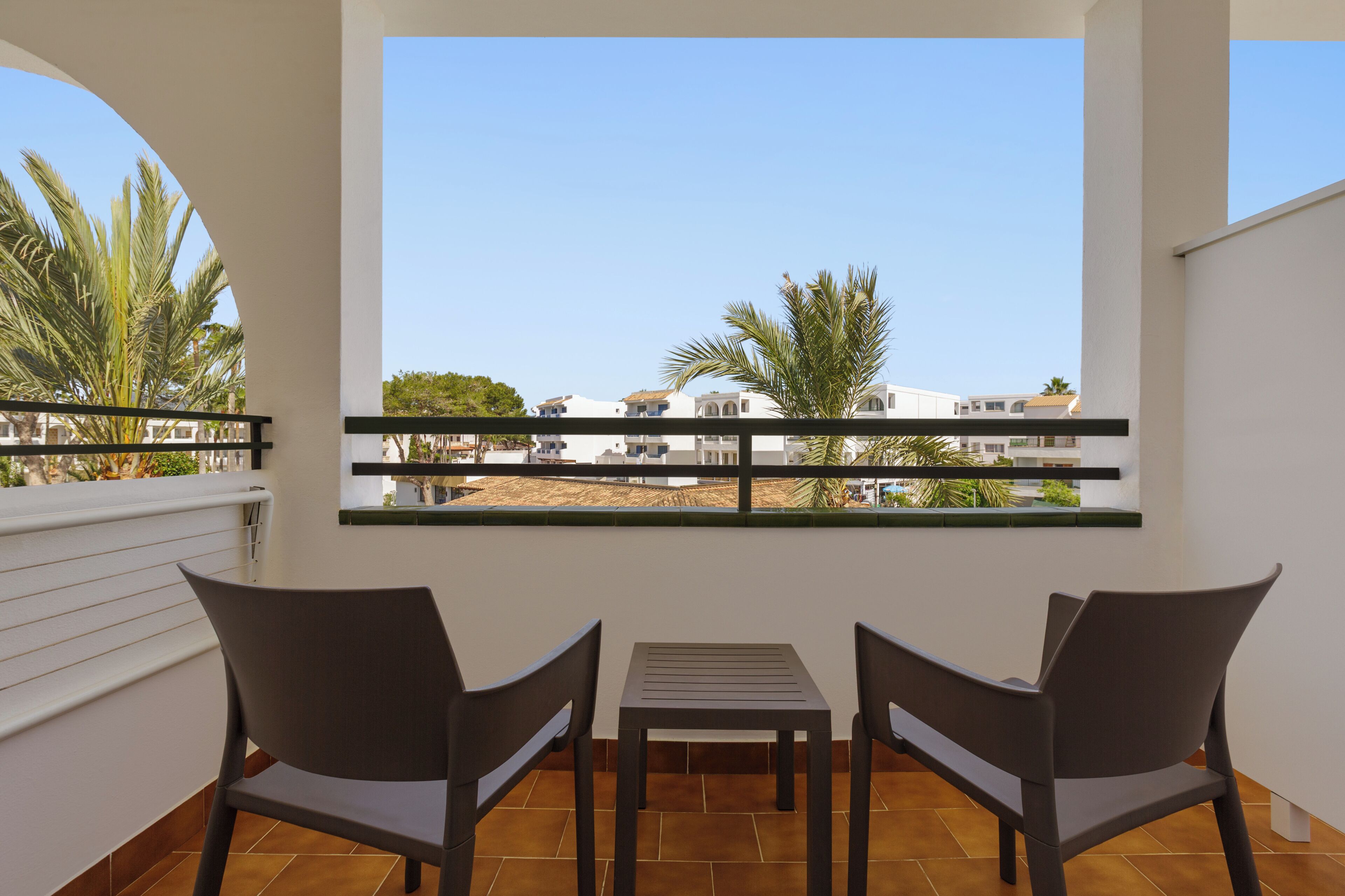 Un balcon avec deux chaises et une table, donnant sur un quartier résidentiel bordé de palmiers et de maisons.