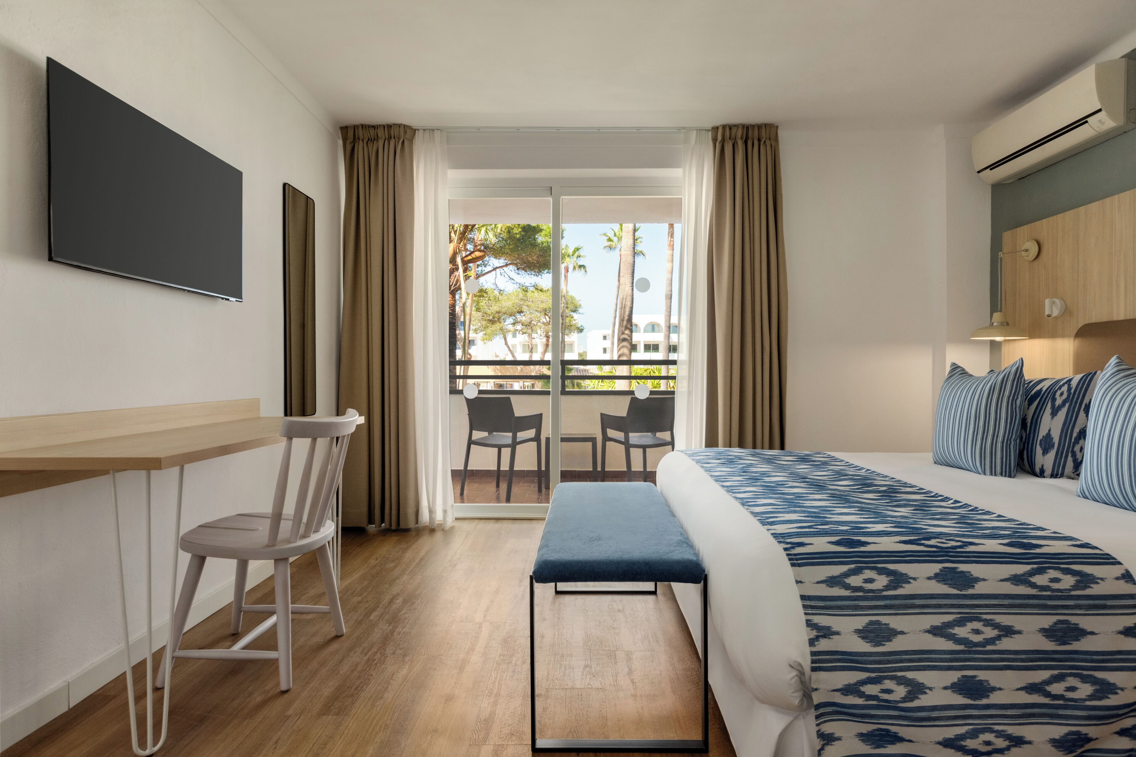 Camera d'albergo con un letto, una scrivania, una sedia, una panca, una TV e un balcone con sedie.
