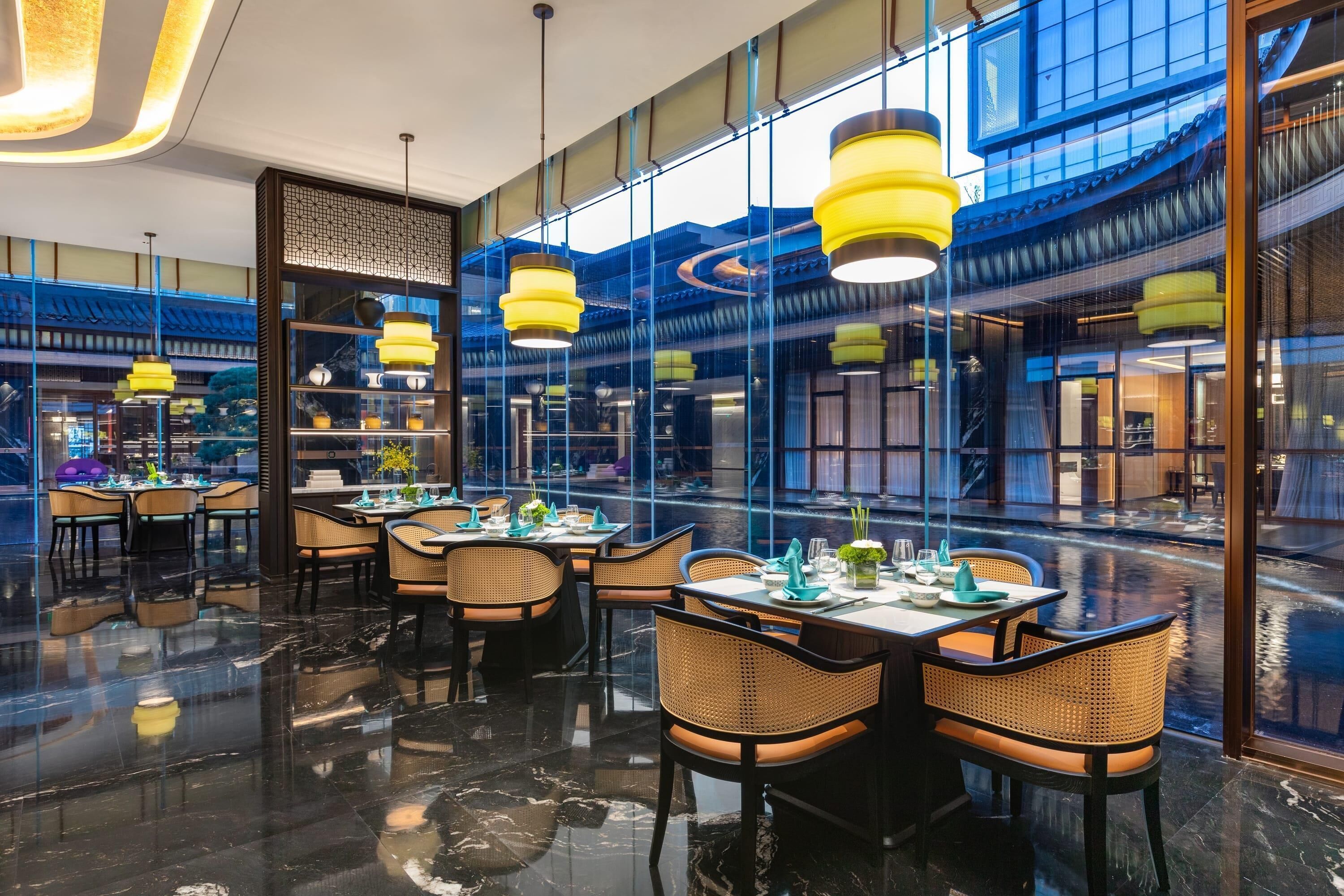 A modern restaurant with a polished floor, yellow pendant lights, and a view of the city through large windows.