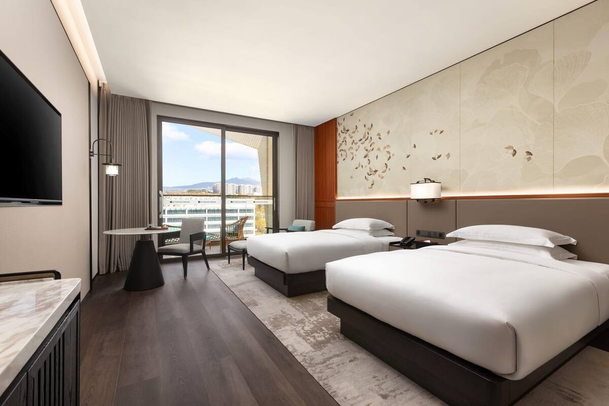 Habitación de hotel con dos camas, un escritorio, una silla, un televisor y un ventanal con vistas a la ciudad.