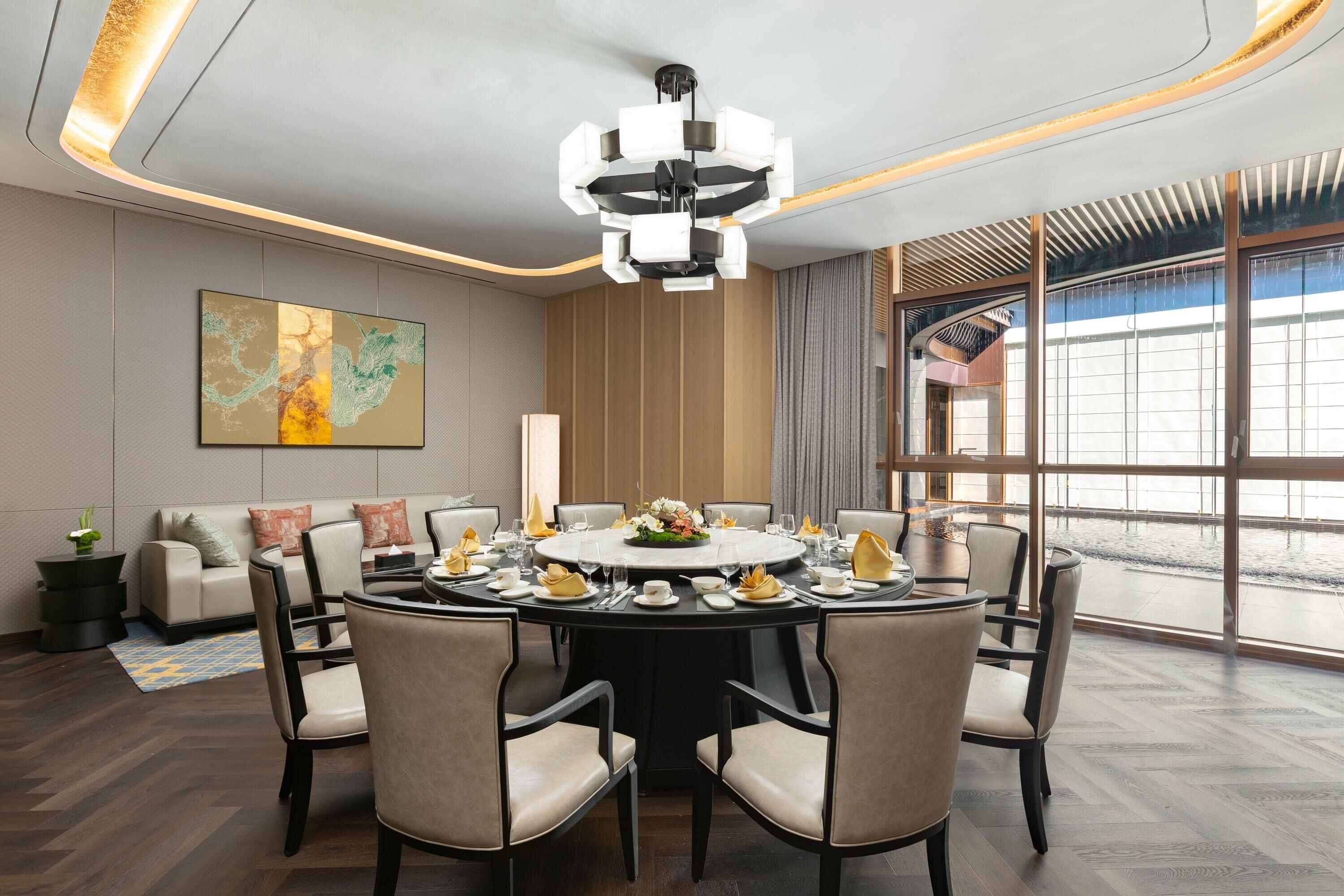 A modern dining area with a round table set for six, surrounded by chairs. A large abstract painting is on the wall, and there is a view of a body of water through large windows.