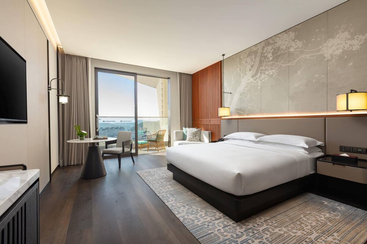 Una habitación de hotel moderna con una cama grande, un televisor de pantalla plana, una zona de comedor con mesa y sillas, y vistas de la ciudad a través de amplios ventanales.
