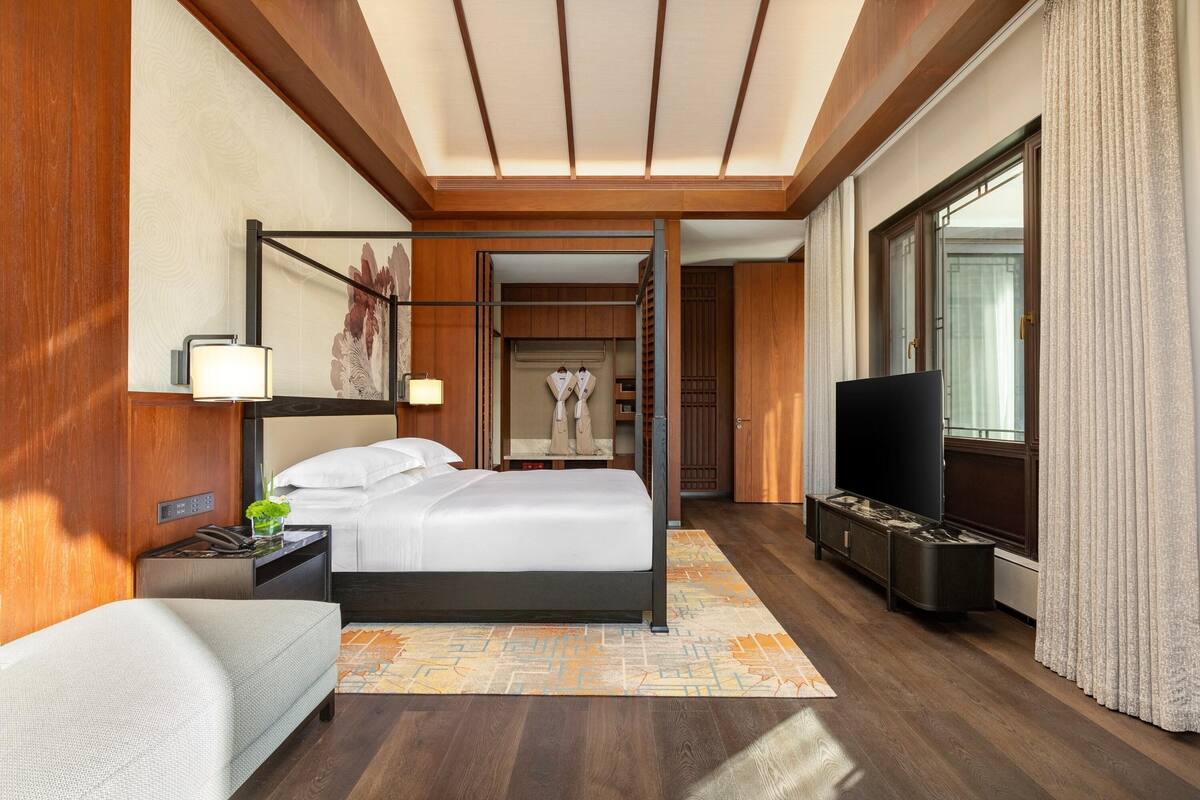 Una habitación de hotel moderna con una cama grande, un televisor, una mesita de noche con lámpara y vistas al pasillo.