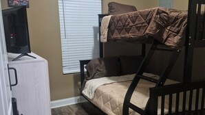 A bunk bed with a quilted mattress, a desk with a computer monitor, and a window with blinds.