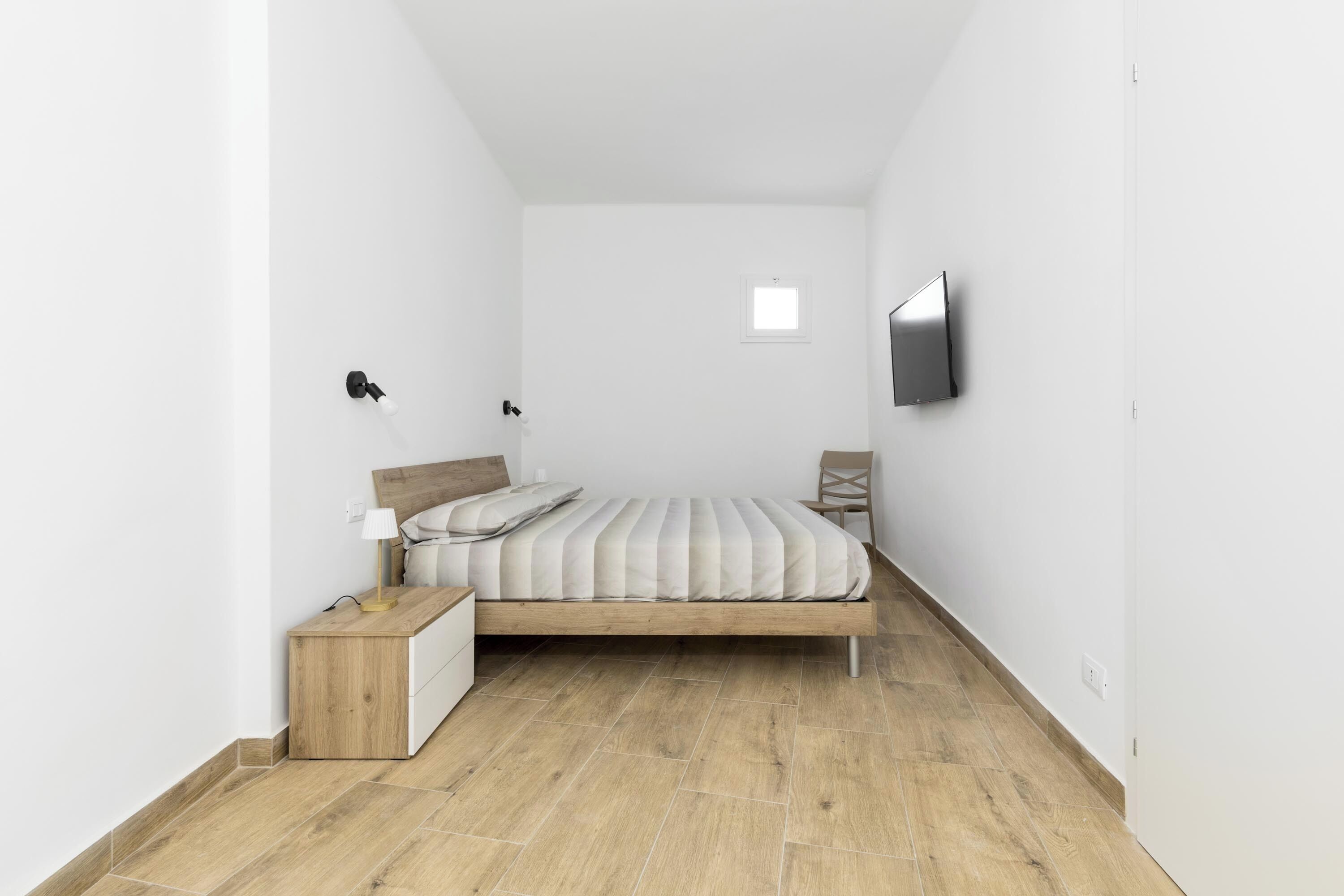 Una camera da letto piccola e minimalista con un letto di legno, un comodino, una sedia e una TV montata a parete.