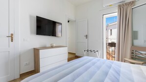 Ein Hotelzimmer mit Bett, einem an der Wand befestigten Fernseher, einer hölzernen Kommode, einem Balkon mit Stuhl und einer Klimaanlage.