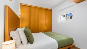 Een slaapkamer met een bed, kussens, een bedlampje, een houten garderobe en een raam met uitzicht.