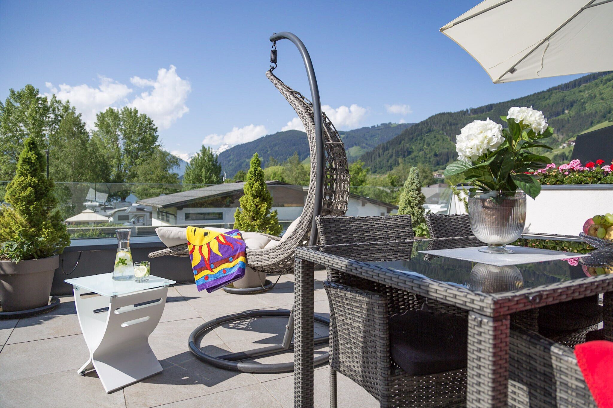An outdoor patio with wicker furniture, a glass table, and a white umbrella. There are potted plants and a view of mountains in the background.