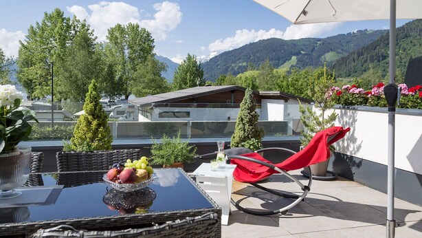 A balcony with wicker furniture, a glass table with fruit, a red chair, and a view of mountains and trees. - Luxury Apartment Silvia, Lake view & Terrace (Zell am See)