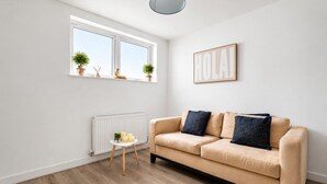 En moderne stue med en beige sofa, et lille hvidt bord med stearinlys, en radiator og et vindue med planter.