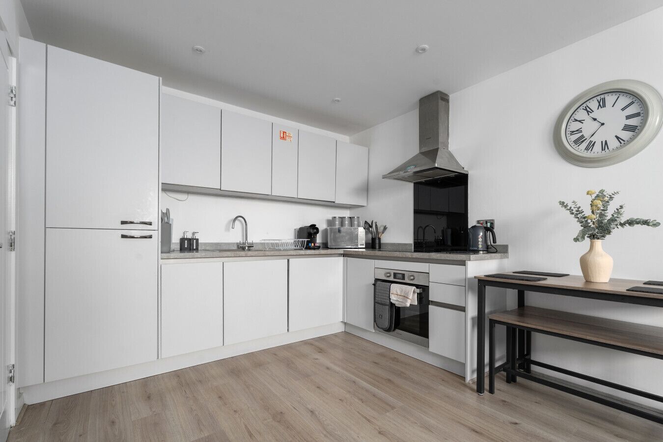 A modern kitchen with white cabinetry, a stainless steel range hood, a built-in oven, a sink, and a clock on the wall.