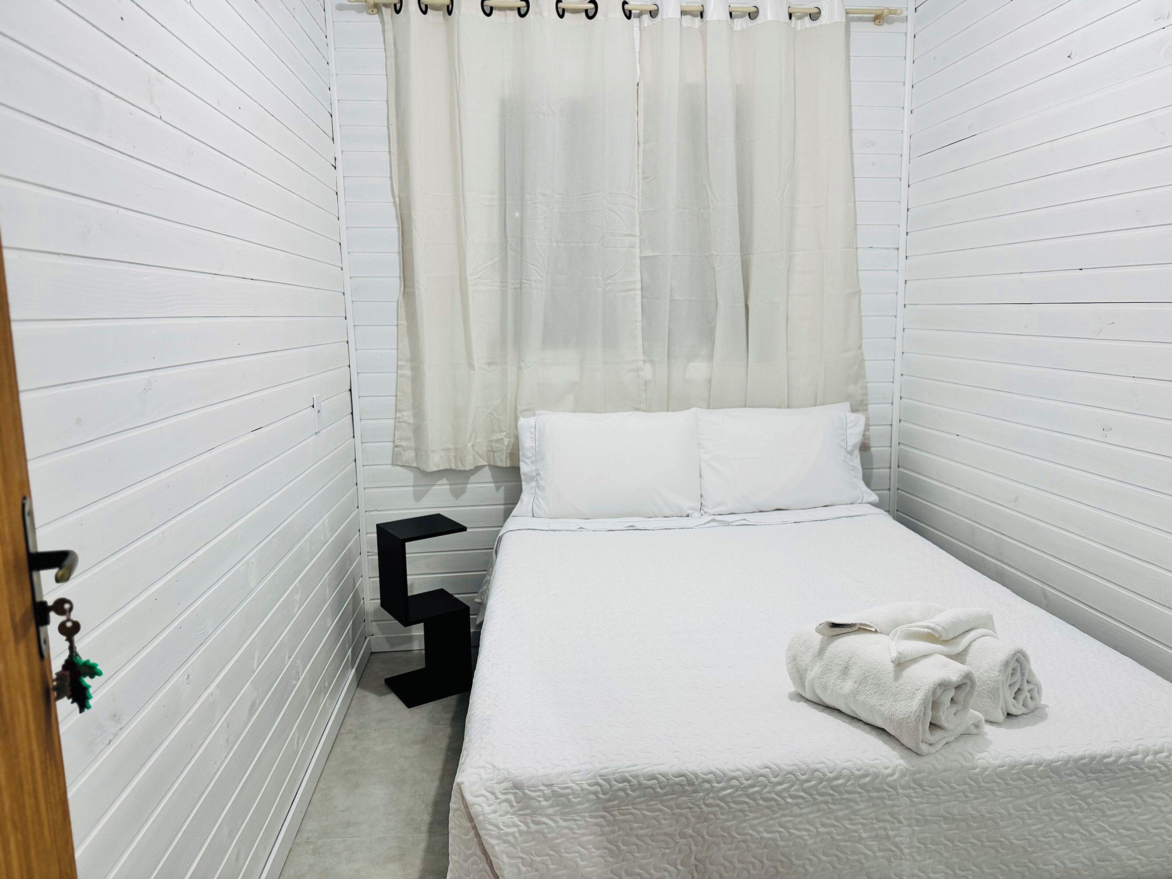A small, white-paneled room with a bed, a black nightstand, and a curtain.