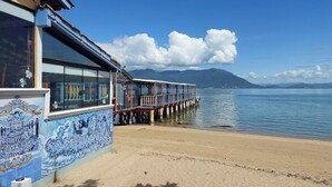 Un bâtiment situé sur la côte, doté d’une façade recouverte de tuiles et d’une jetée s’avançant dans des eaux calmes, avec des montagnes en arrière-plan.
