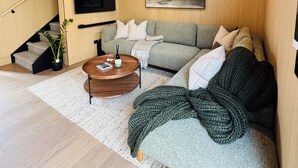 A modern living room with a wooden ceiling, a flat-screen TV mounted on the wall, a sofa with cushions, a coffee table, and a staircase with a plant. - Luxury villa & carpark, retreat close to everything (Queenstown)
