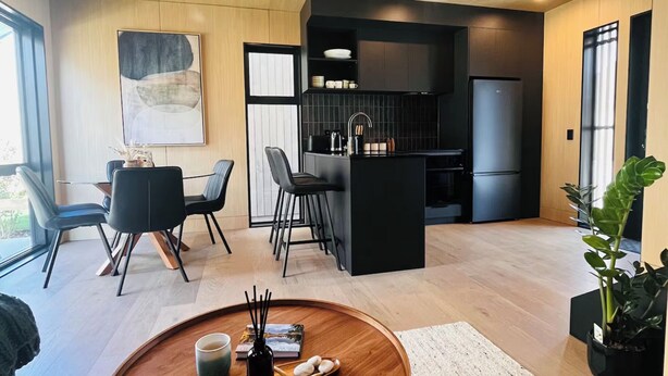 A modern living room with a round wooden coffee table, a dining area with chairs, a kitchen with black cabinetry, and a large window providing natural light. - Luxury villa & carpark, retreat close to everything (Queenstown)
