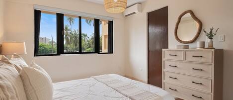 A bedroom with a bed, a dresser, a window with a view of palm trees, and a hanging light fixture.