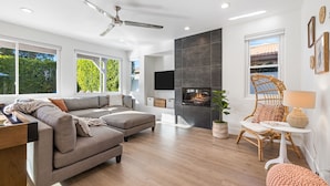 A modern living room with a sofa, a fireplace, a ceiling fan, and a large window. - Wren by Nest Villas | Private Pool, Spa & Firepit (Indio)