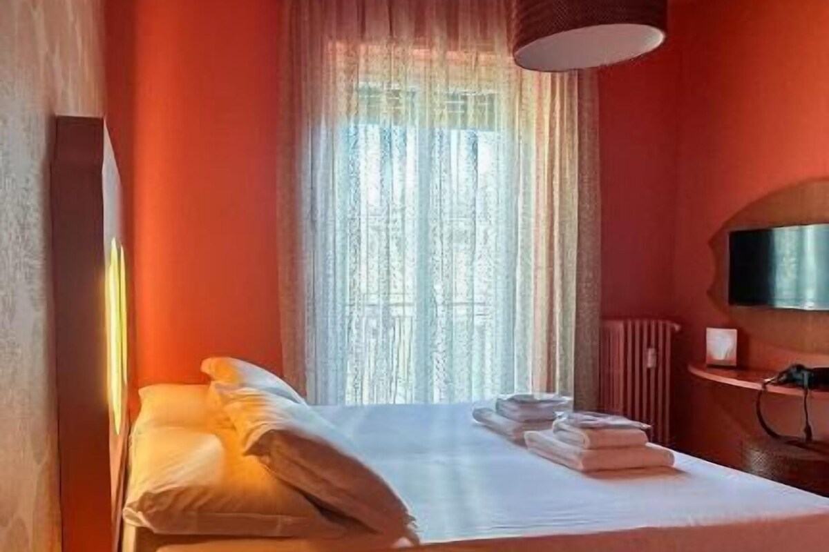 Un llit individual amb una taula de nit i una làmpada, una finestra amb cortines translúcides i un televisor fixat a la paret.
