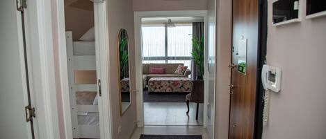 A hallway leading to a bedroom with a bed, a dresser, and a mirror. There is a door on the right and a window with curtains in the background.