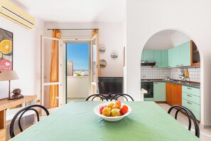 Un coin repas avec une table dressée pour deux personnes, un saladier rempli de fruits et une vue sur la ville par une porte ouverte.