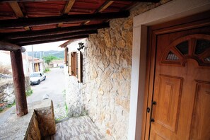 Edificio de piedra con puerta y contraventanas de madera, un acceso empedrado y vista a una calle con un automóvil estacionado.