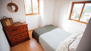 Une chambre à coucher avec une commode en bois, un lit recouvert d’une couvre-lit à rayures et deux fenêtres laissant entrer la lumière naturelle.