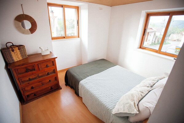 Un dormitorio con un aparador de madera, una cama con colcha a rayas y dos ventanas que dejan entrar luz natural.