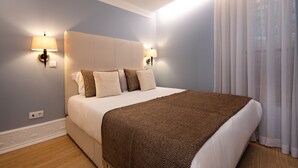 Un letto rifatto con cura, decorato con una coperta fantasia, due lampade a parete e una finestra con tende leggere.