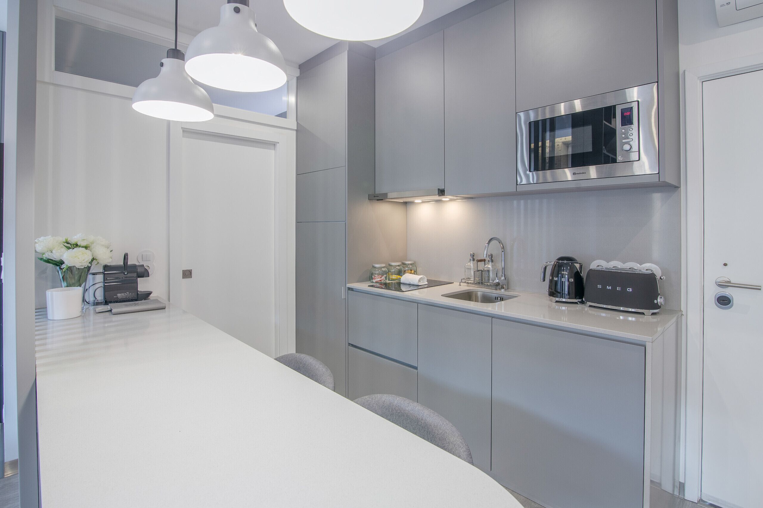 Uma cozinha moderna com micro-ondas embutido, pia e torradeira. Há luminárias pendentes sobre o balcão e um vaso com flores à esquerda.